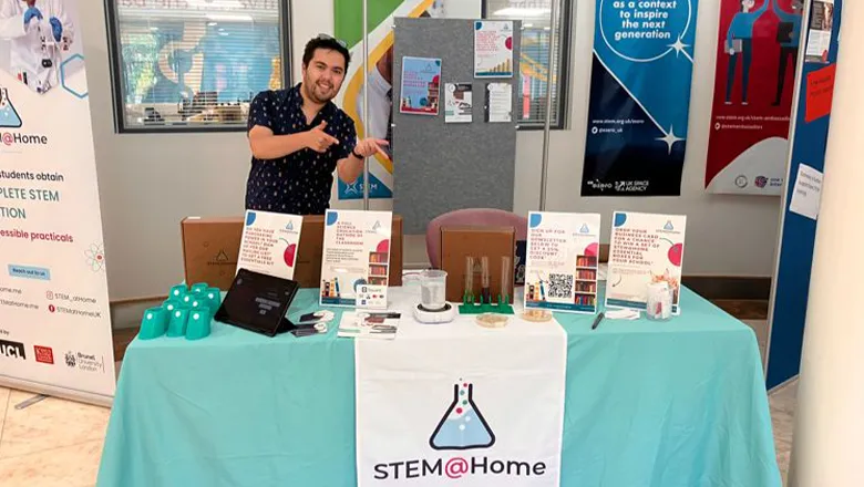 Francis is smiling and standing behind a stall upon which information about STEM@Home can be seen.