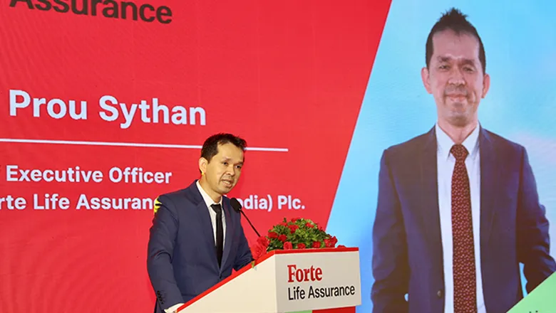 Sythan is pictured speaking at a conference. He is wearing a dark blue suit and a black tie, and he is delivering his speech from a lectern.