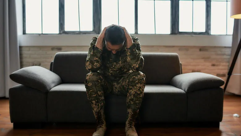 Man in military uniform sat on a sofa with his head in his hands