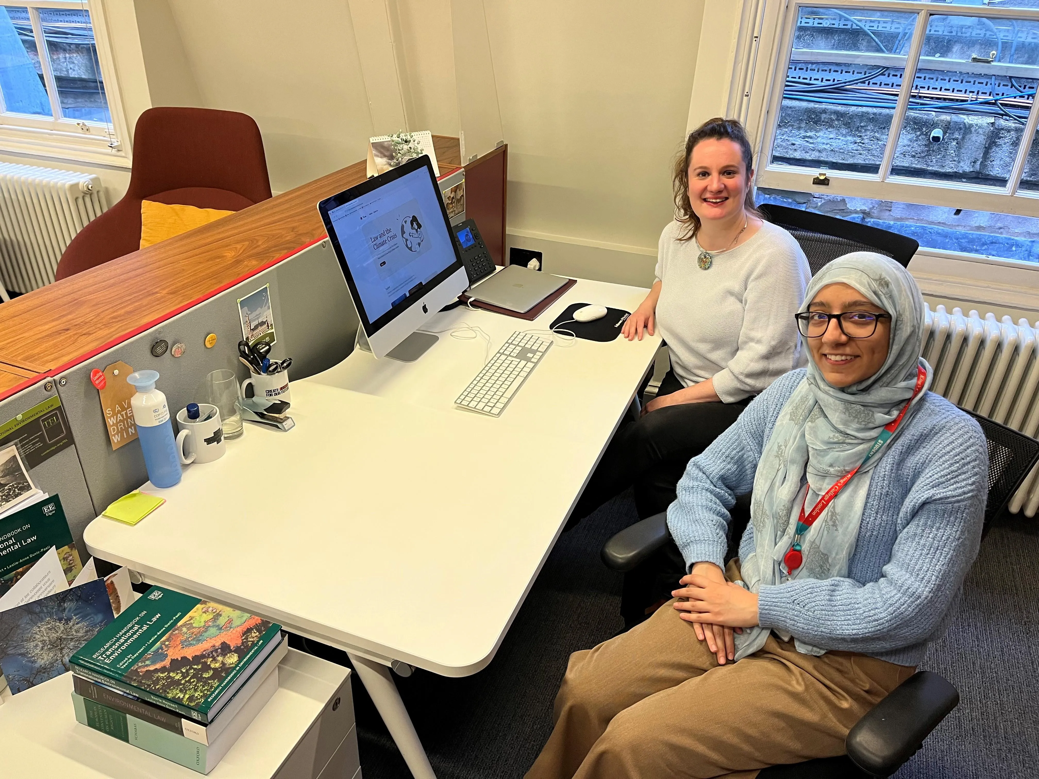 Two smiling women at a desk in front of a computer. One is in blue jumper with a hijab and the other is in a white sweater with a necklace. 