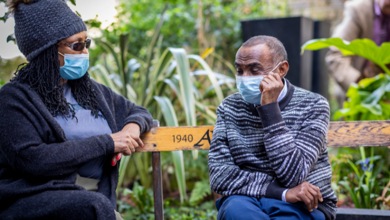 Two older people sit in a garden conversing, both wearing blue surgical face masks.