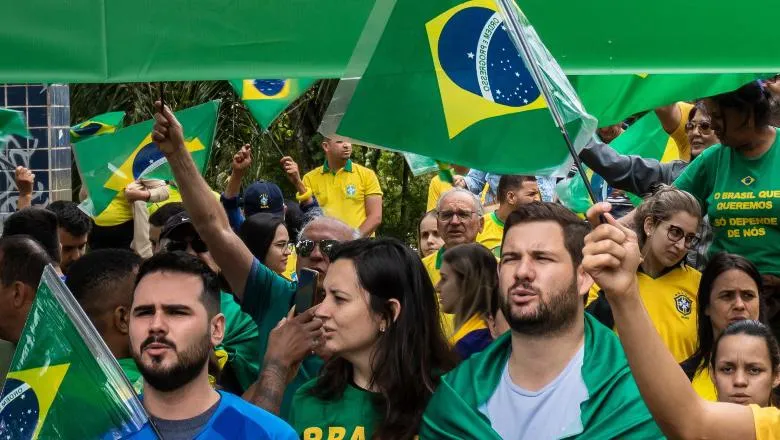 Protesters waving the Brazilian flag, with three men in the foreground holding their hand over their heart and singing.