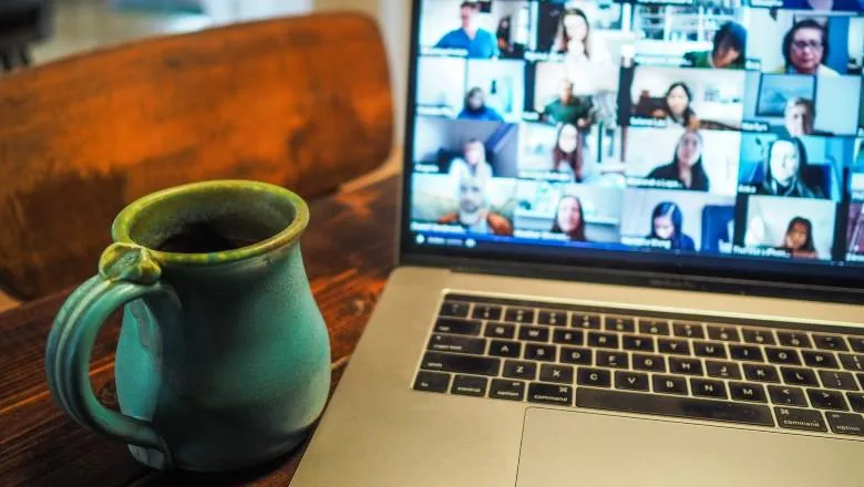 Photo of a laptop on which there is a Zoom session going on with about 20-30 people. Next to the laptop is a blue mug.