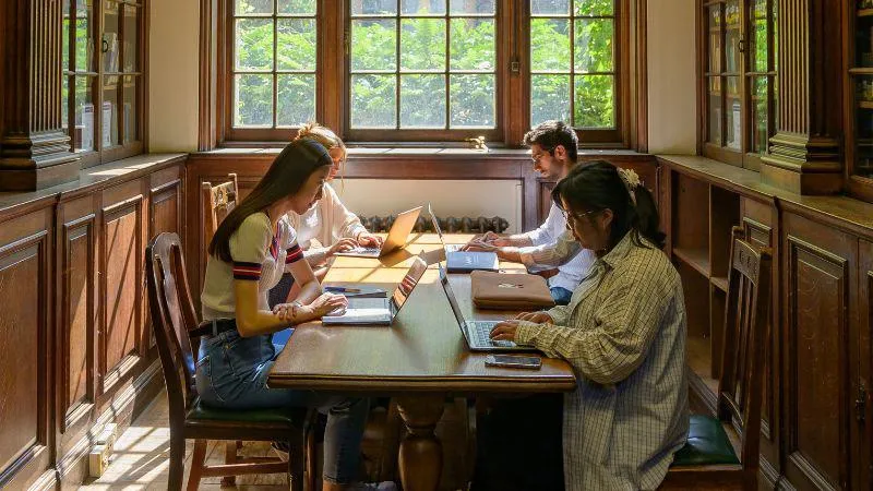 Four students sit at a wooden table in the library, each student has a laptop in front of them.