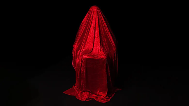 A woman sat on a chair, draped in a red velvet cloth against a black background