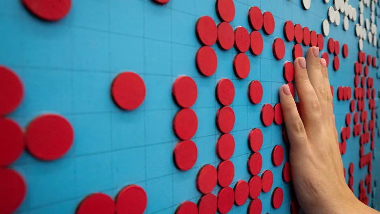 A hand is exploring a work of art made of large braille text that has been cut from painted wood. The braille is arranged in bands: a lower band of red on a bright blue background, and an upper band of white on a bright blue background. 