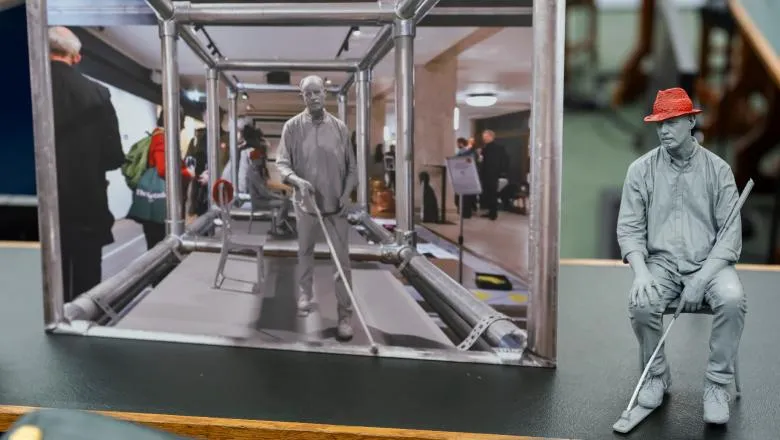 A model of a man with a red had sat on a chair with a white cane next to a photo of a similar figure