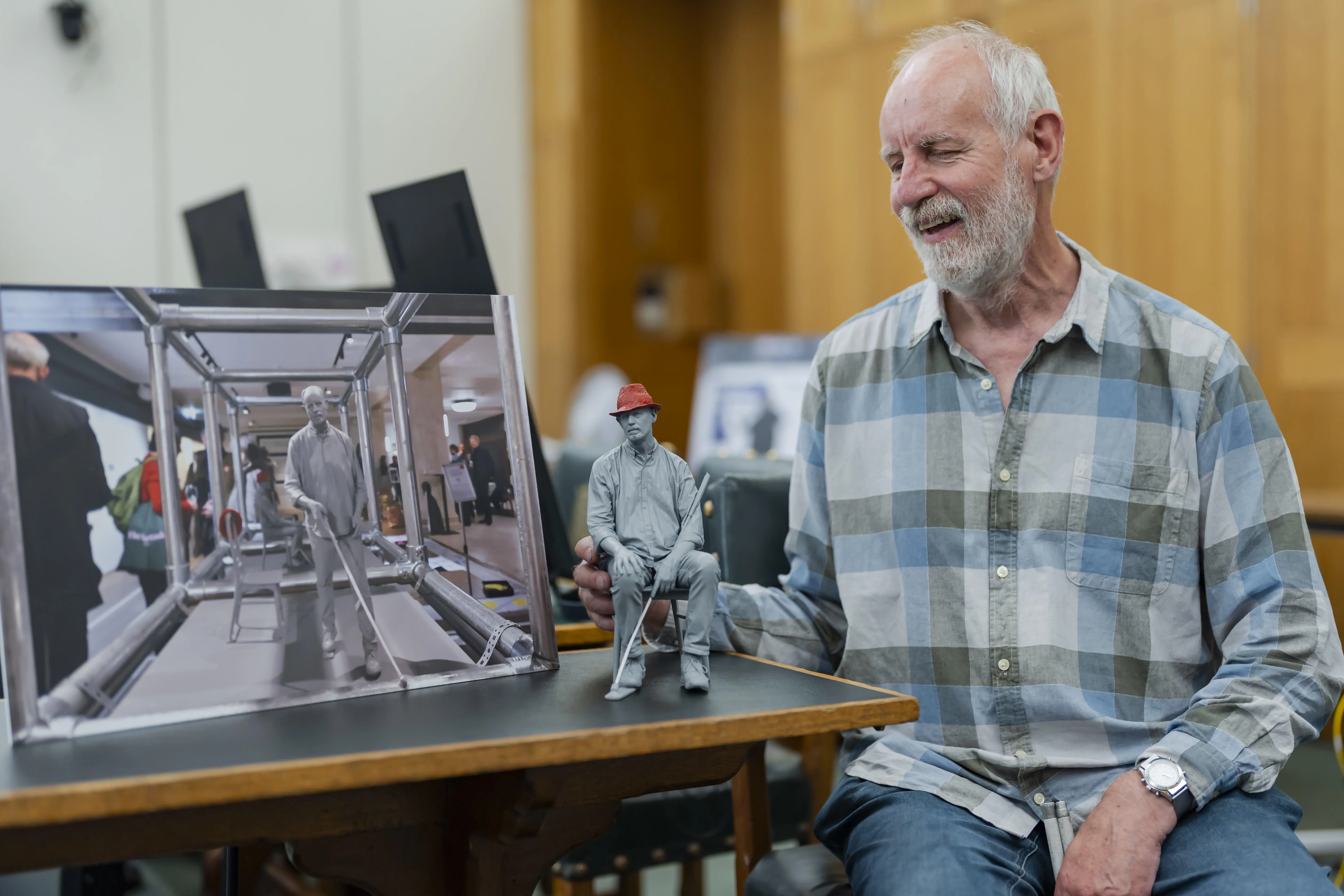 Artist David Johnson smiles beside a small sculptural self-portrait and a photo of his full exhibited work