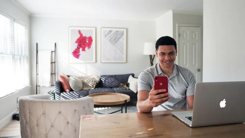 a man sits at his laptop in a bright living room, he is smiling at the phone he is holding