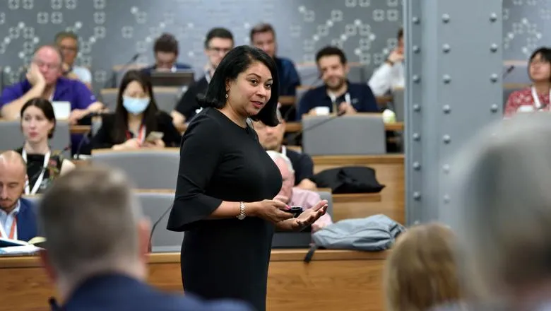 An image of Maura Scott, wearing a black dress, speaking to an audience sitting in a lecture theatre