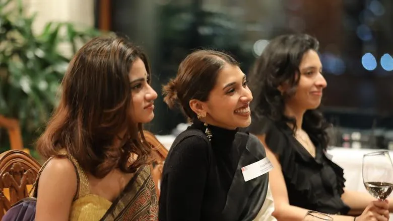 Three women, two in sarees, smiling while listening to a speaker