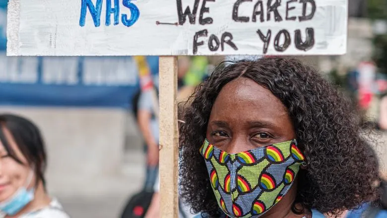 A black nurse wearing a face covering carries a placard saying 'We cared for you'