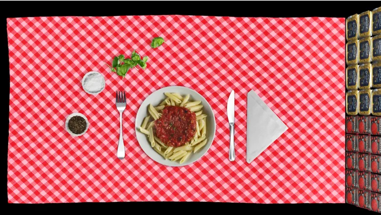 a view from above of the ingredients of a tomato pasta dish, laid out on a checkered table cloth. On the right of the image are stacks of pasta packets and tin cans.