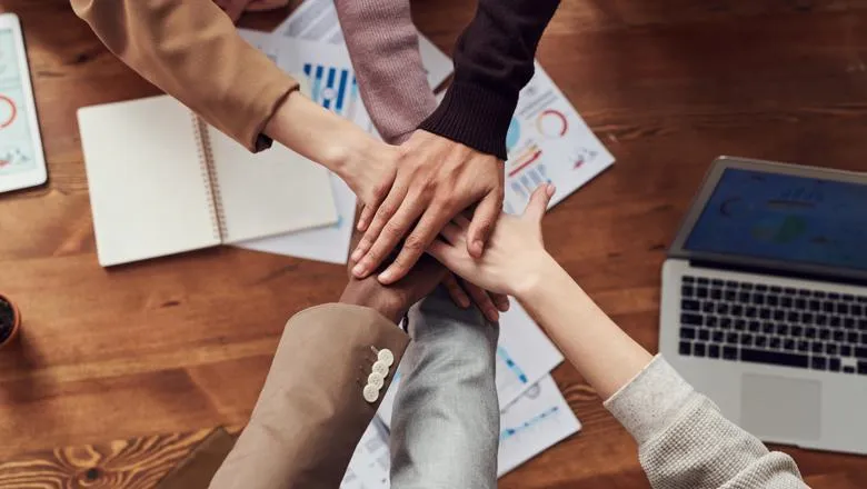 Six hands layered on top of each other above a work desk in a gesture celebrating the team