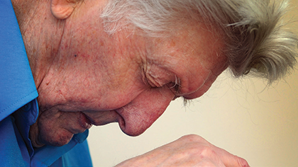 Seated elderly man leaning on walking stick bowing his head with his eys closed