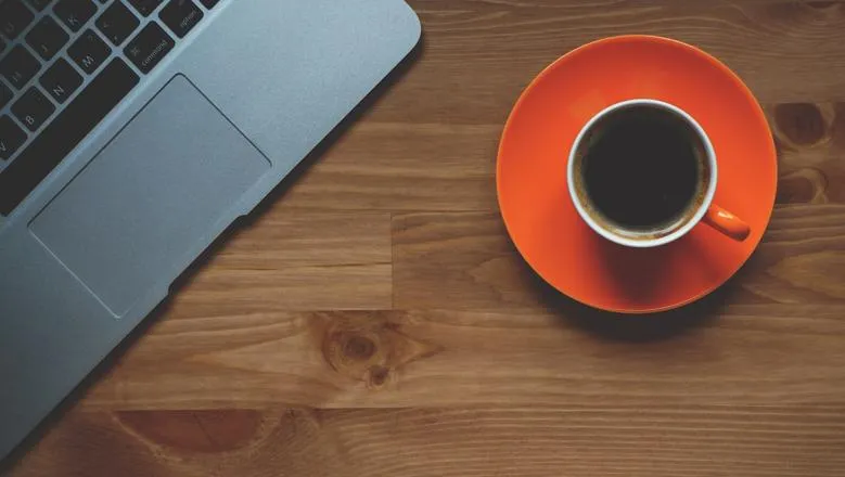 Black coffee in orange cup with saucer on a wooden table with laptop