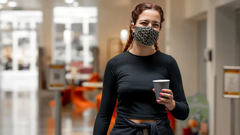A student walking down a corridor wearing a mask and carrying a coffee.