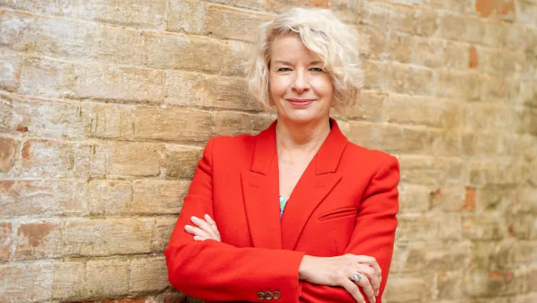 Tania Rhodes-Taylor stands in front of a brick wall, arms folded and smiling, wearing a red blazer