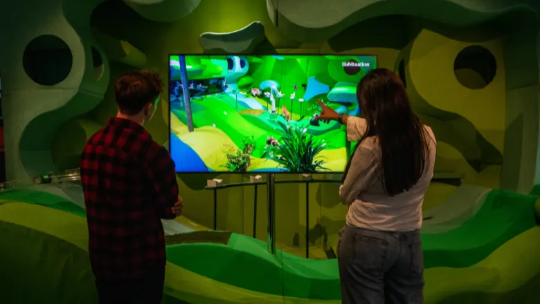 Two figures stare at a television screen in the centre of the shot, their backs to camera. They are looking at a bright fluorescent green screen. One of the figures is pointing, but the image of the screen is hard to make out. In the background, green swirls and shapes can be seen. 