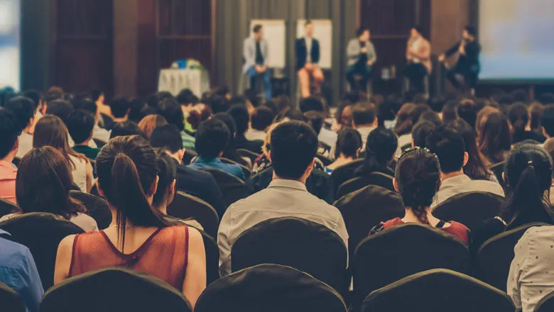 An audience from behind, facing a stage where speakers are talking.