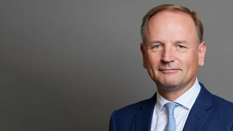 Simon Stevens wears a blue suit with white shirt and blue tie. He is photographed in front of a grey backdrop.