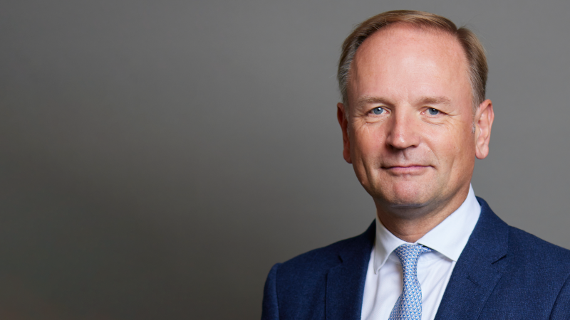 Simon Stevens wears a blue suit with white shirt and blue tie. He is photographed in front of a grey backdrop.