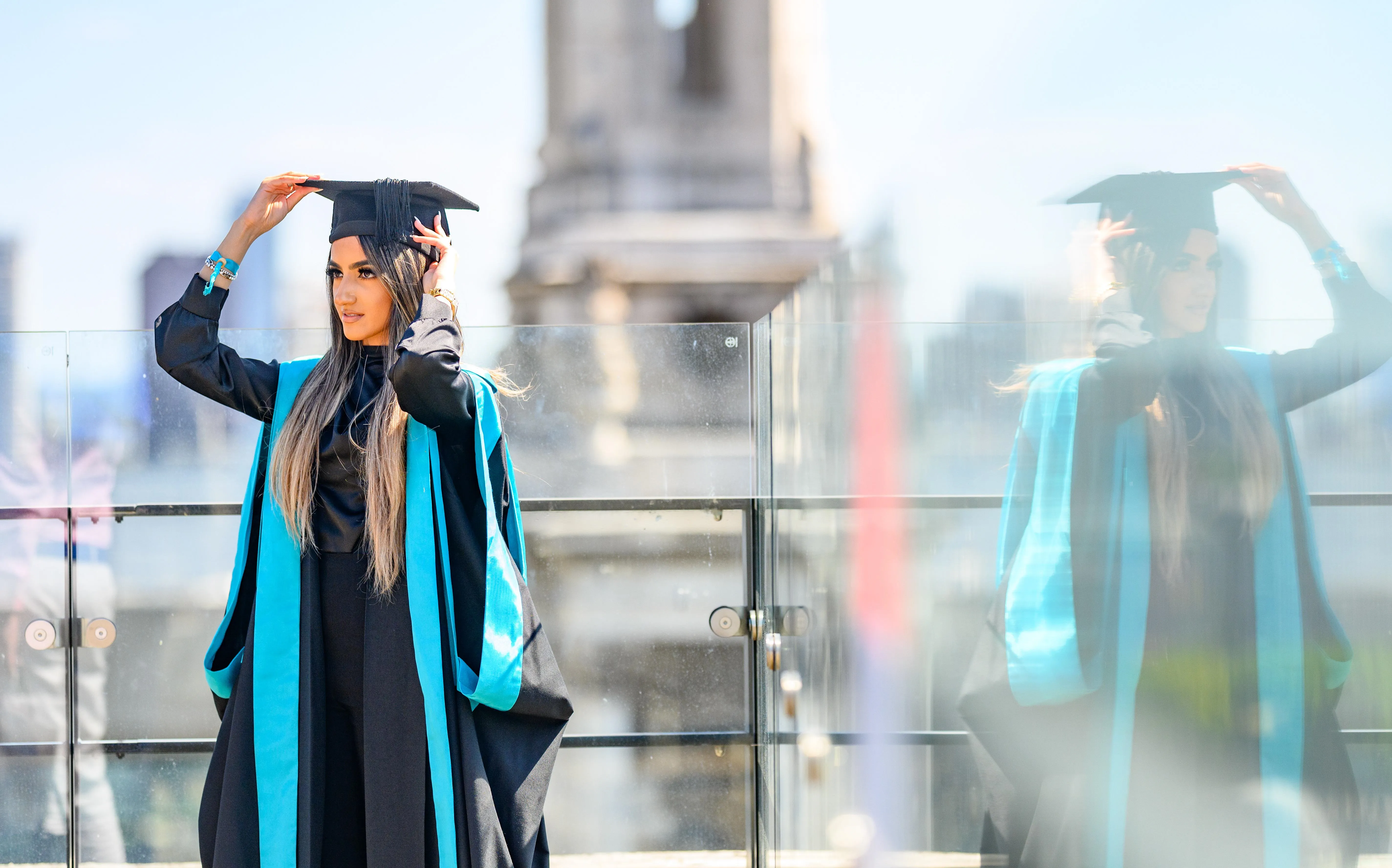 A King's Business School graduate wearing academic dress adjusts her mortar board. Her image is reflected in the glass railing to her side, and we can see the London skyline behind her.
