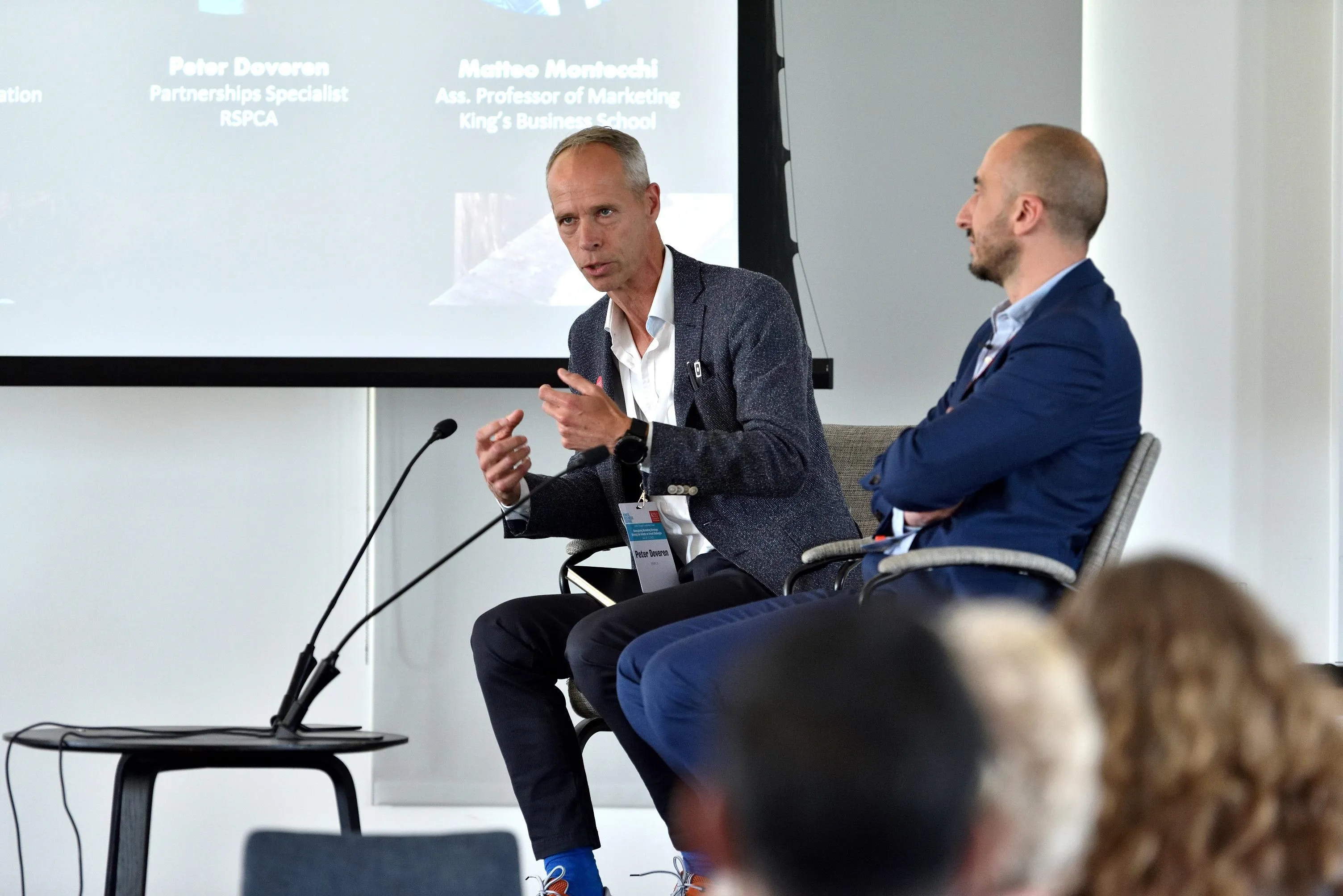 Two men are seated in front of an audience at an event. They are engaged in lively conversation and one gestures with his hands as he leans towards a microphone.