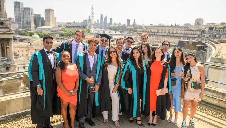 A group of smiling King's Business School graduates pose in their graduation gowns on a sunny roof terrace. We can see the London Eye and Houses of Parliament in the background.