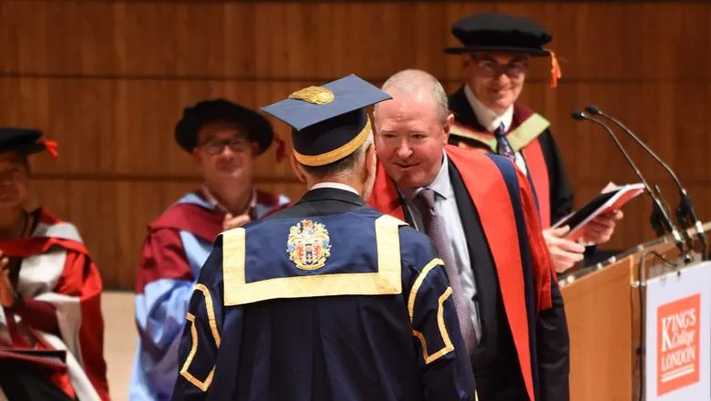 A man, Warwick Hunt, wearing red academic robes, shakes hands with a senior representative of King's (facing away from us) at a degree ceremony on the stage at the Royal Festival Hall. Other representatives of King's, also wearing academic dress, are sitting in the background.