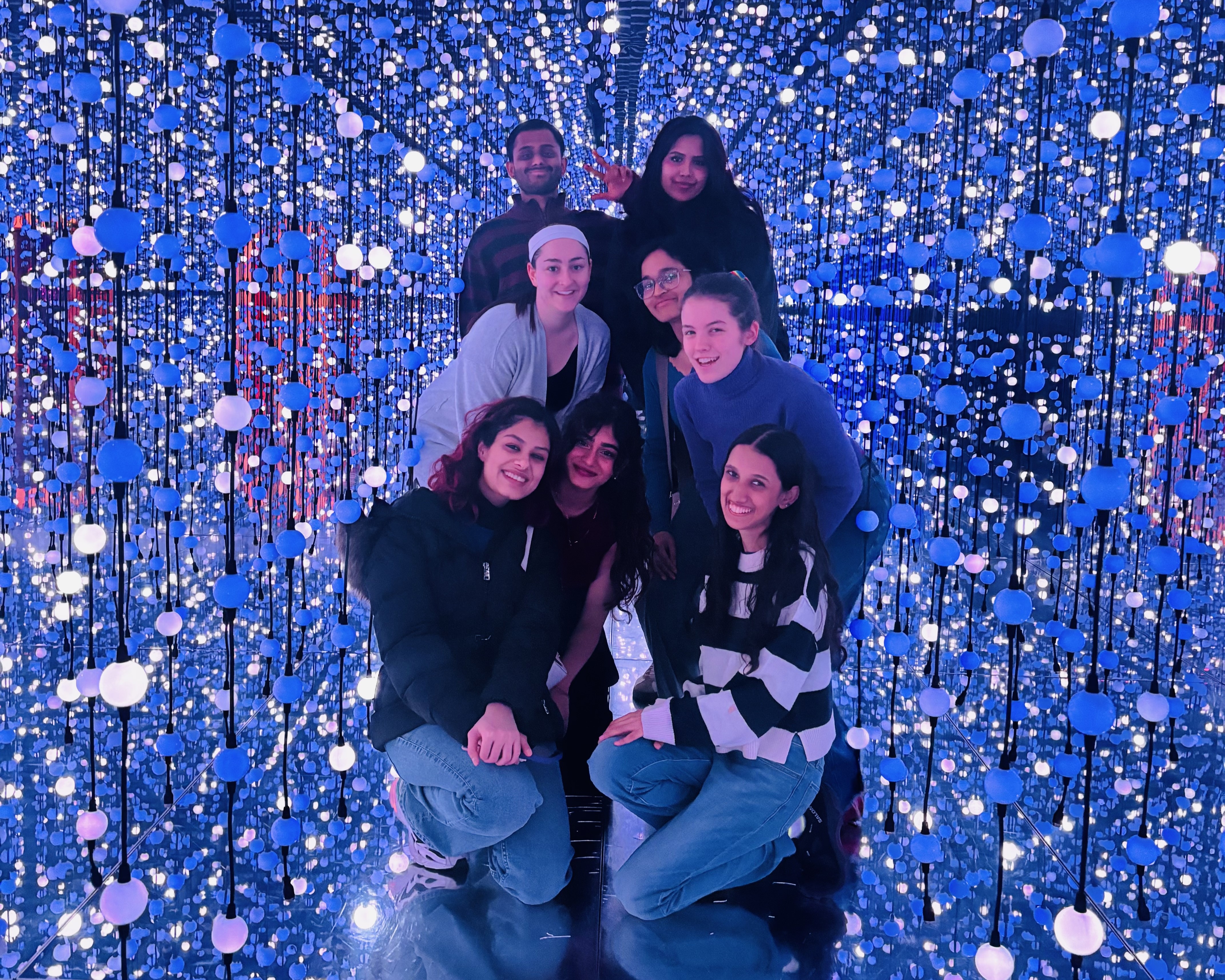 A group of students posing together inside an immersive mirrored room filled with blue and white lights suspended at different heights. The group is gathered closely in the center, smiling at the camera, surrounded by the glowing installation.