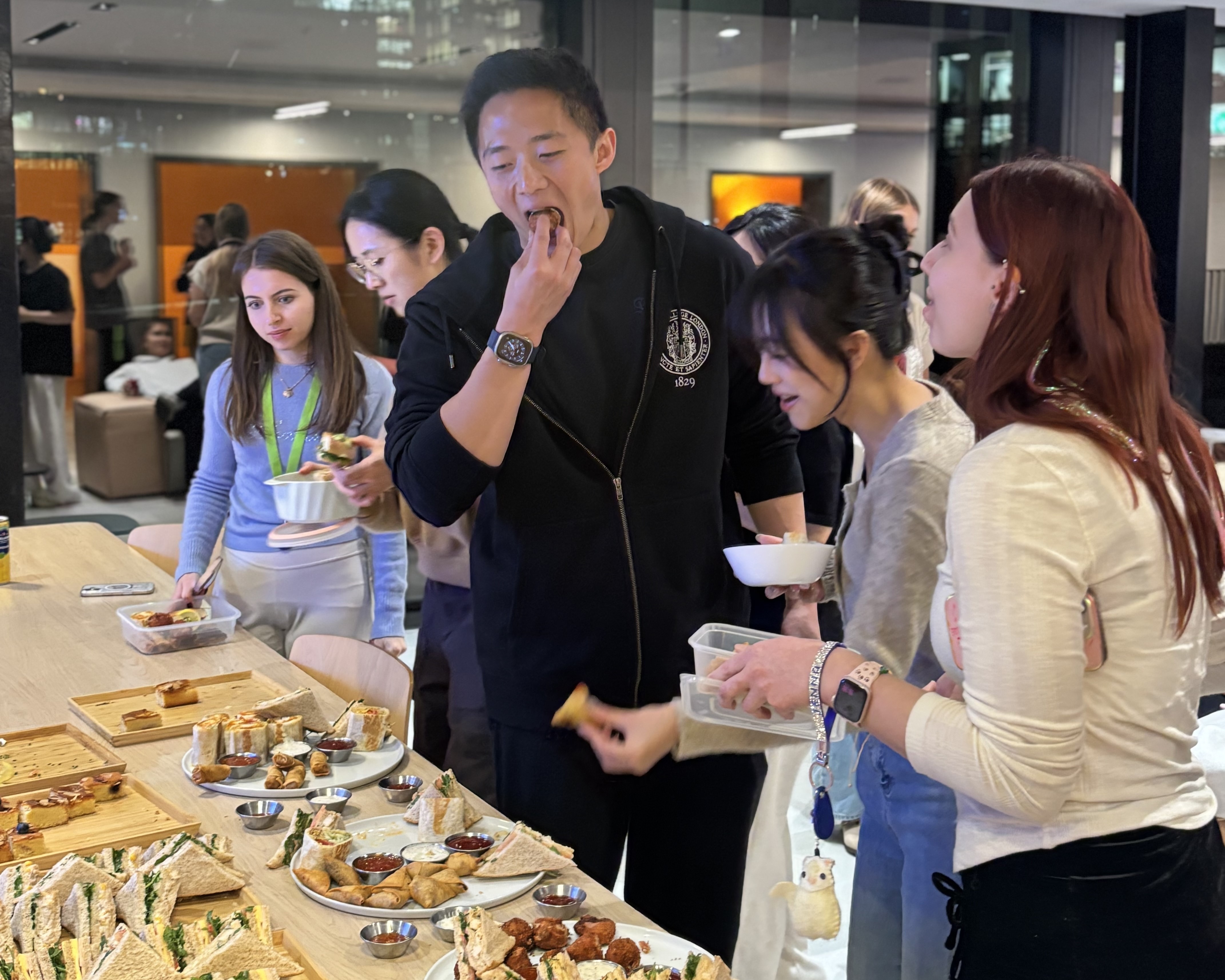 People standing around a long table indoors, selecting food from trays filled with sandwiches and small dishes. The group is gathered in a bright, modern room while interacting near the buffet-style spread.