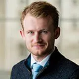 Dr James Corke-Webster looking directly at camera with short hair, wearing a blue tie, light shirt and dark jumper infront of a blurred background.