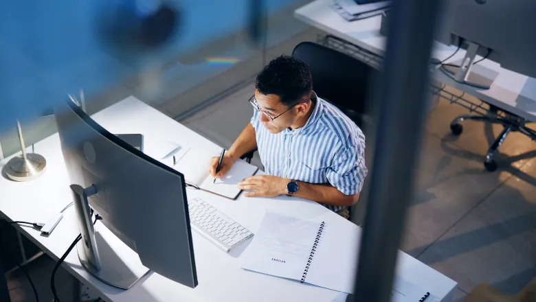 A researcher writing at a desk and looking at a computer screen