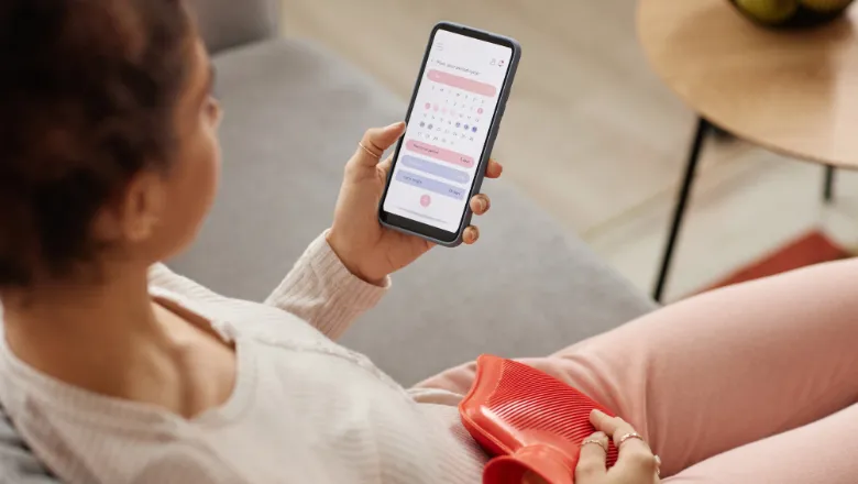 High angle closeup of young woman holding smartphone with period tracker app on screen and holding hot water bottle on belly