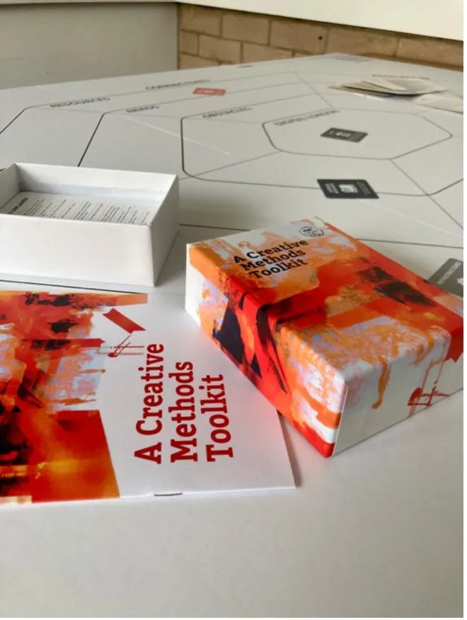 Orange board game box in the foreground on a white table with the white boardgame in the background looking from above. 