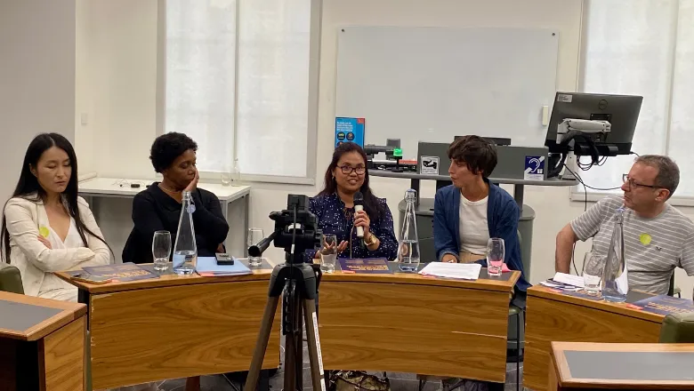 The image shows five panel members sitting at the table. A woman in the middle is speaking to the audience.