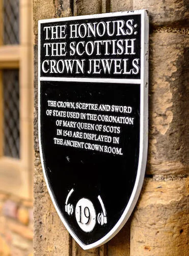 The image shows a metal plate on the wall saying: 'The Honours: The Scottish Crown Jewels. The crown, sceptre and sword of state used in the coronation of Mary Queen of Scots in 1543 are displayed in the ancient Crown Room.