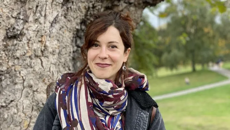 A portrait of a woman standing in front of a tree and green landscape. Her hair is tied up. She is lightly smiling. She is wearing a dark grey jacket and a scarf.