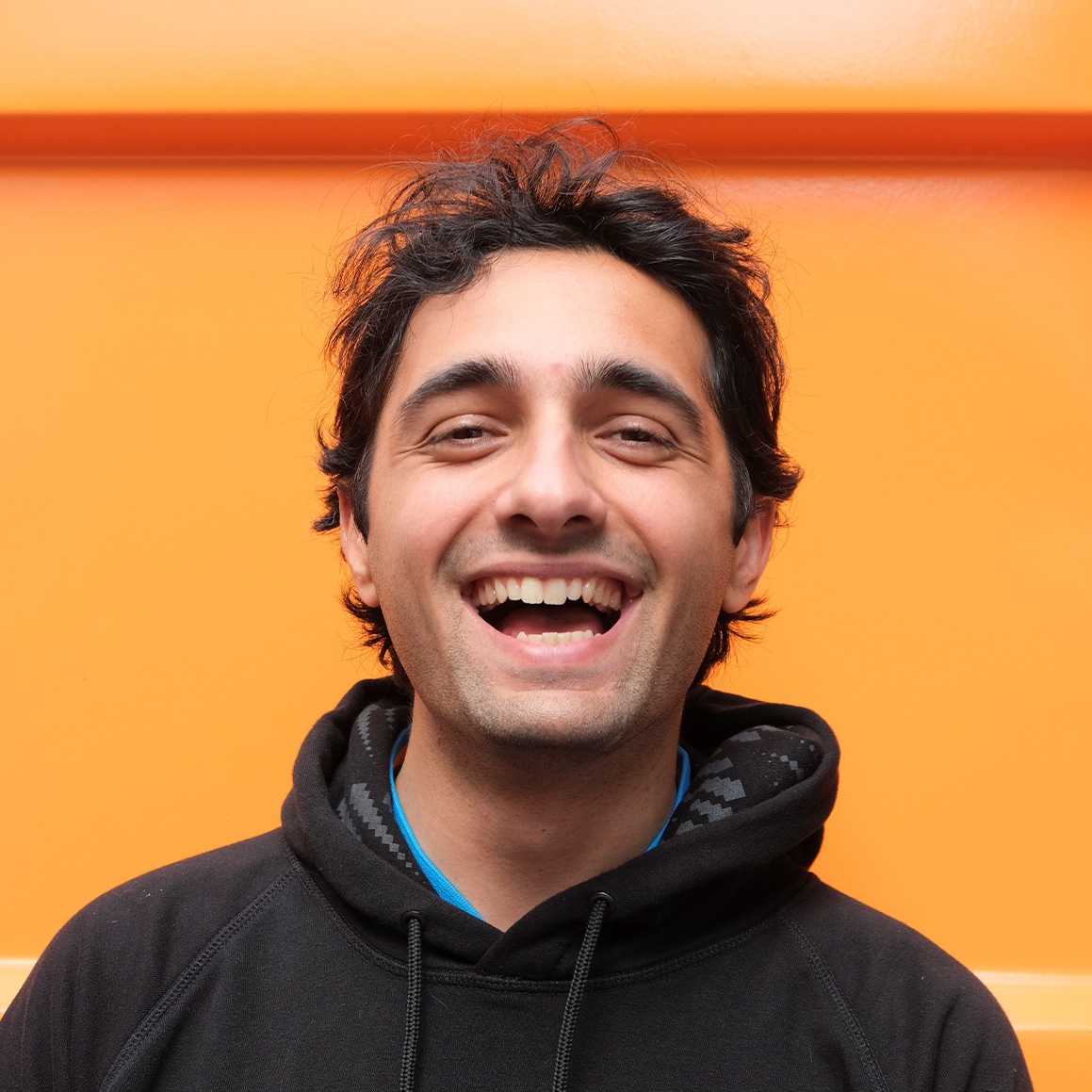 Alex Webb, a young person with dark and curly short hair, smiling, while wearing a black hoodie.