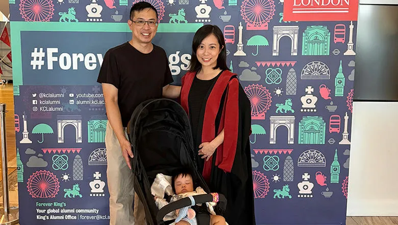 A young woman with dark hair and wearing graduation robes stands next to a man with dark hair, glasses, a black t-shirt and tan trousers. In front of them is a buggy with a child inside. In the background is a banner that reads ‘King’s College London’. 