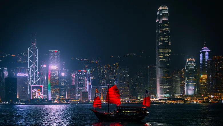The Hong Kong skyline at night, with a boat with red sails in the foreground.