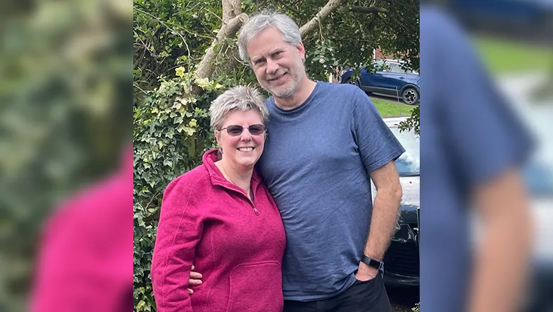 Ann and Keith are stood side by side, smiling. They are stood in front of a tree.