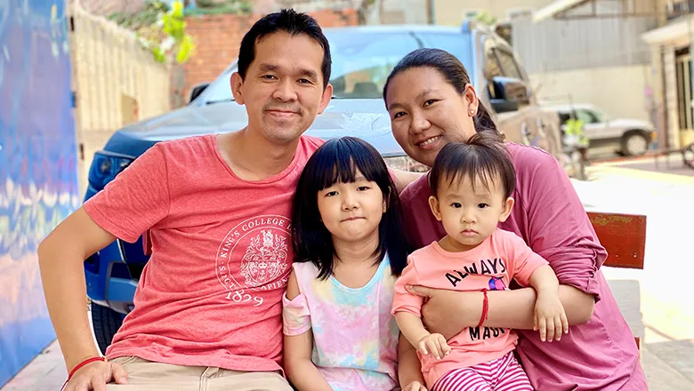 Sythan Prou is pictured with his wife and two daughters on a bench. He is wearing a red t-shirt which has the King's College London logo emblazoned on it.