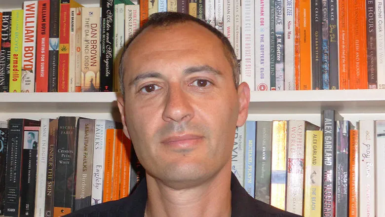 A head and shoulders photo of a man with short, cropped hair wearing a black shirt. In the background is a bookshelf with lots of books.