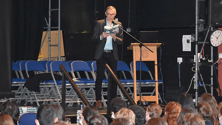 A man with short hair wearing a light blue shirt, a dark jacket and dark trousers stands on stage. He is reading a book to an audience of schoolchildren.