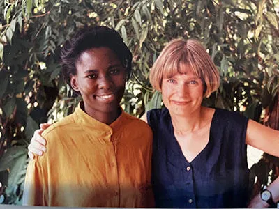 A woman with dark hair wearing a yellow top stands next to another young woman with short blonde hair wearing a blue top. They are outside in a hot climate. Both women smile at the camera.