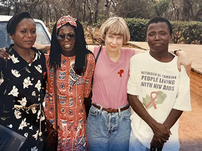 A group of four women stand arm-in-arm. On the left is a women with short, dark hair wearing a black and white dress. Next to her is a woman wearing glasses and an orange dress with a flower design and long dark hair. Next to her is to a woman with short blonde hair wearing blue jeans and a pink top. Next to her is a woman with short dark hair wearing a t-shirt that bears the logo ‘Network of Zambian people living with HIV and Aids’. The stand outside in a hot climate