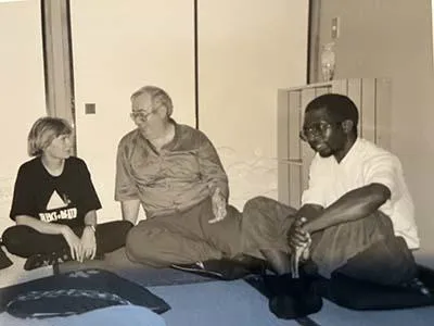A black-and-whiter photo showing three people sitting on the floor in a room. On the left is a woman with short blonde hair wearing a dark t-shirt. Next to her is a middle-aged man with glasses wearing a light-coloured short. Next to him is a man with short cropped hair and glasses wearing a white shirt. All three look to be deep in conversation