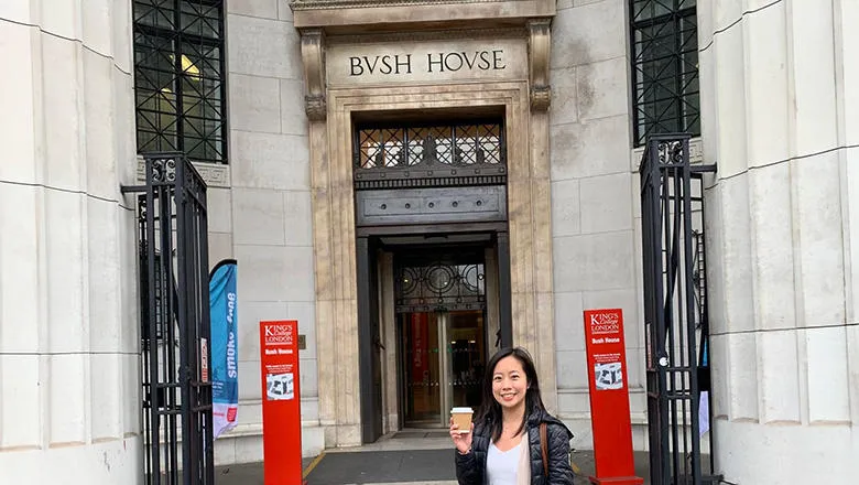 A young woman with dark hair and wearing a black, coat, black trousers and a white top stands outside the ornate main entrance to Bush House on the Strand. She smiles and holds a cup of coffee in one hand. 